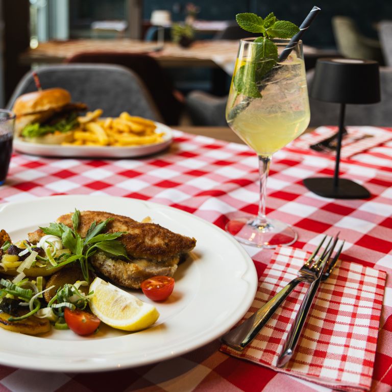 Ein paniertes Schnitzel, serviert mit gebratenen Kartoffeln, Lauch, Zucchini, Kirschtomaten und einer Zitronenspalte auf einem weißen Teller.