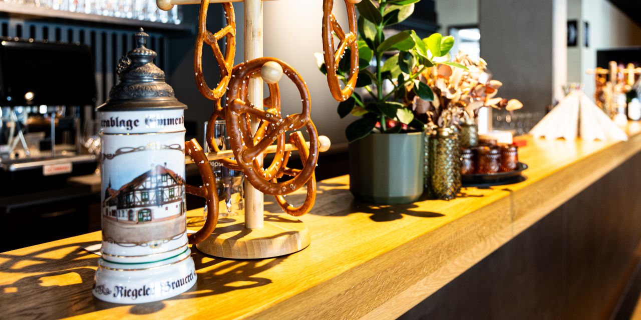Hölzerne Theke, darauf ein traditioneller Bierkrug mit Zinndeckel und ein hölzerner Brezenständer mit aufgehängten Brezen. Daneben eine grüne Pflanze in einem Topf.